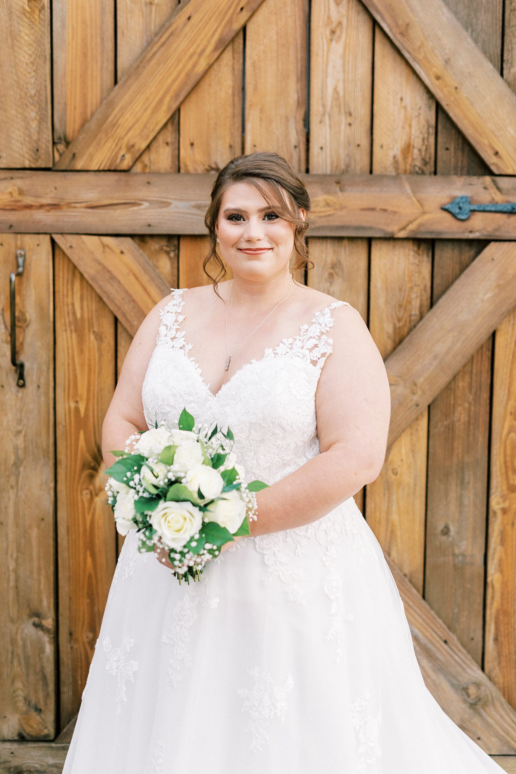bride in lace wedding gown holds bouquet in front wooden barn doors