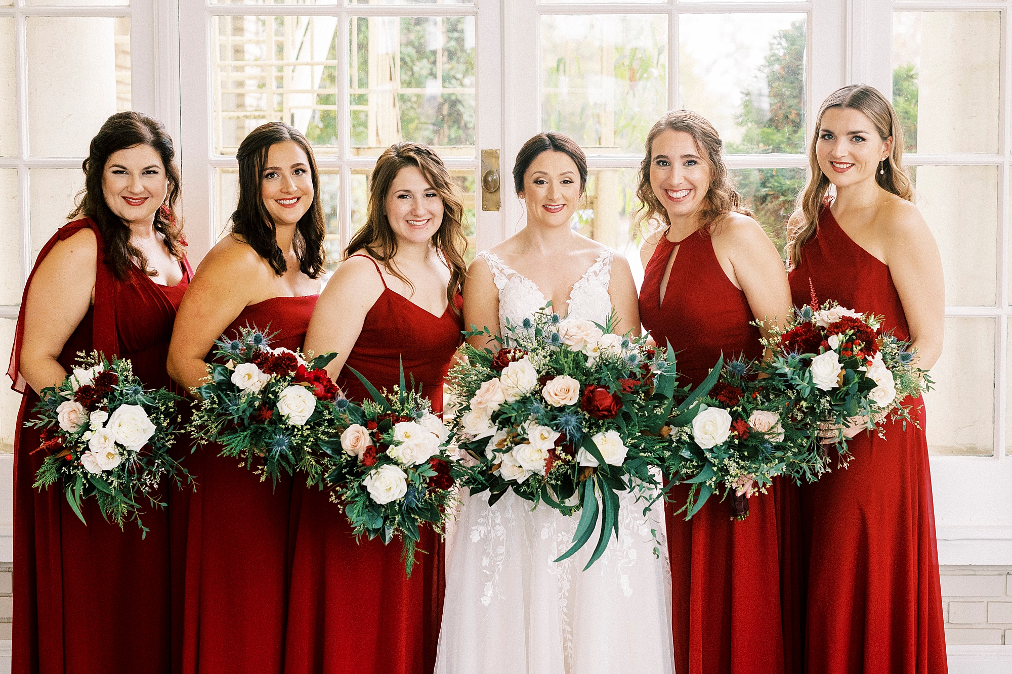 bride poses with bridesmaids in red gowns with red and white bouquets for classic winter wedding at Separk Mansion