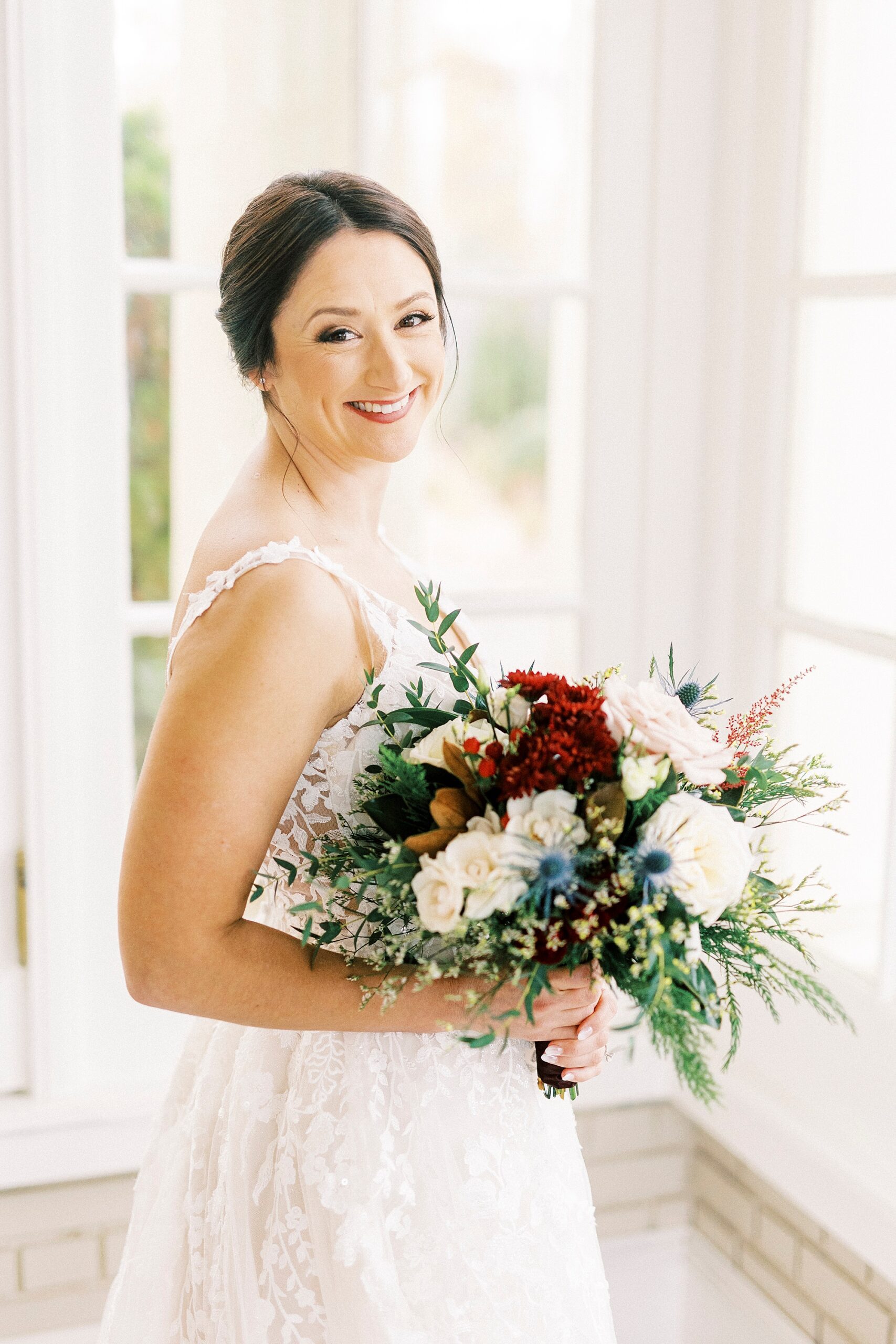 bride holds bouquet of white and red flowers smiling over her shoulder