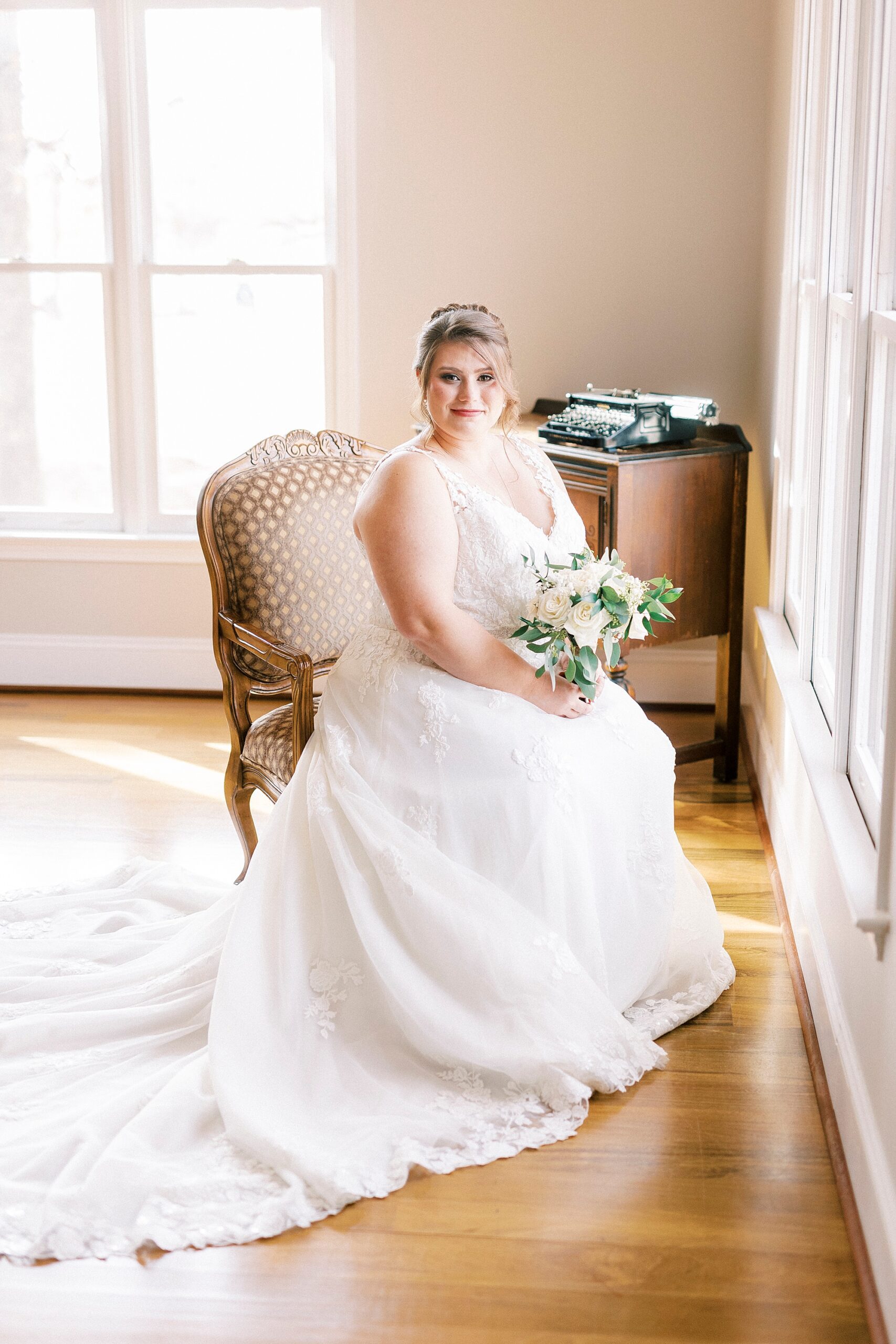 bride sits in tan chair at the Farm at Brusharbor in bridal suite with bouquet of white flowers 