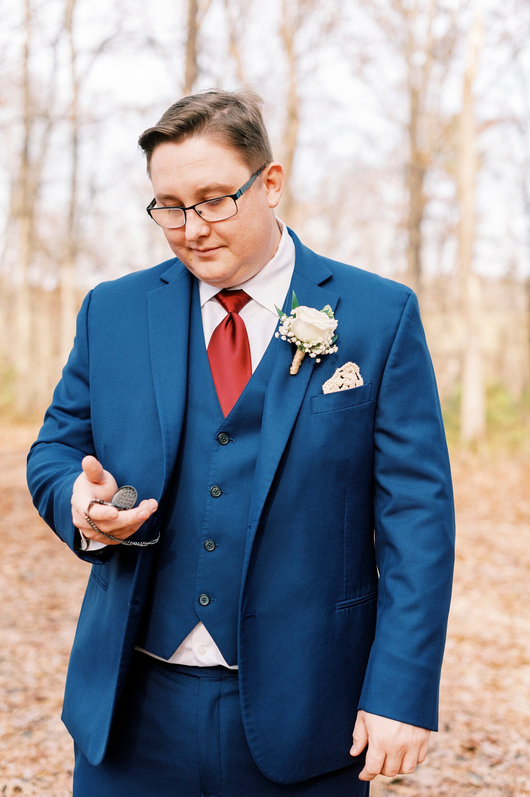 groom in navy suit with red tie looks at custom pocket watch 