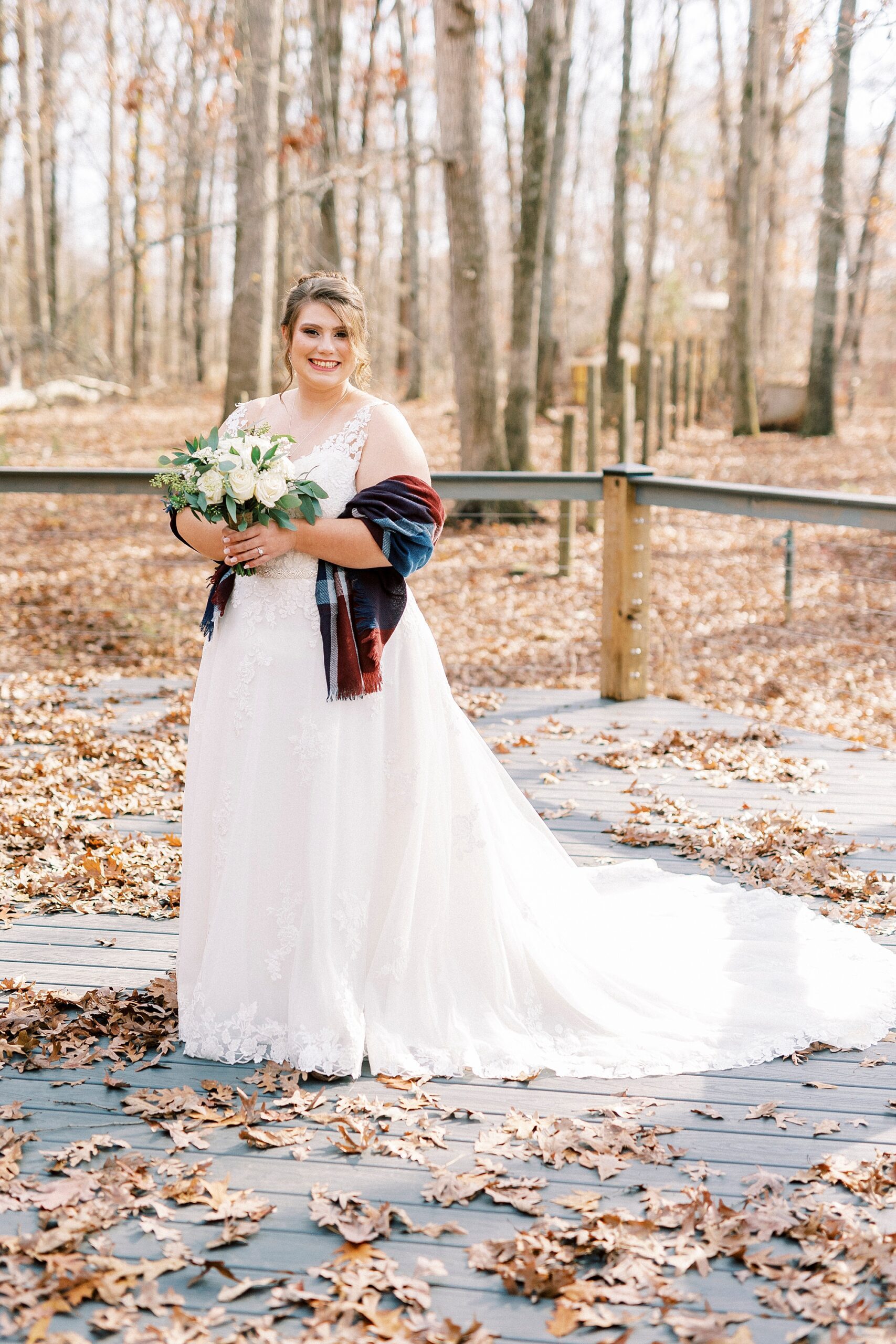 bride stands on fallen leaves wearing red plaid shawl around her wedding dress
