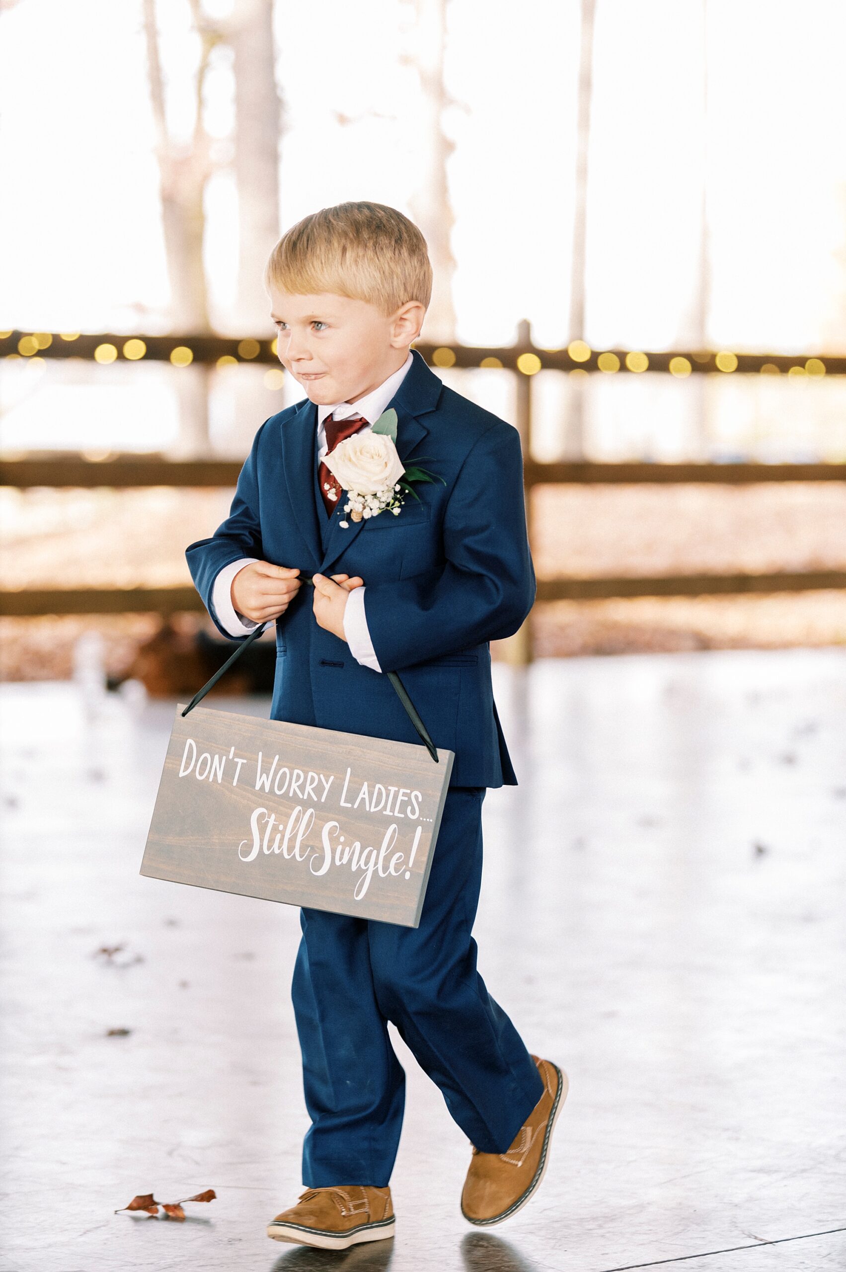 ring bearer in navy suit walks down aisle for ceremony with wooden sign at the Farm at Brusharbor