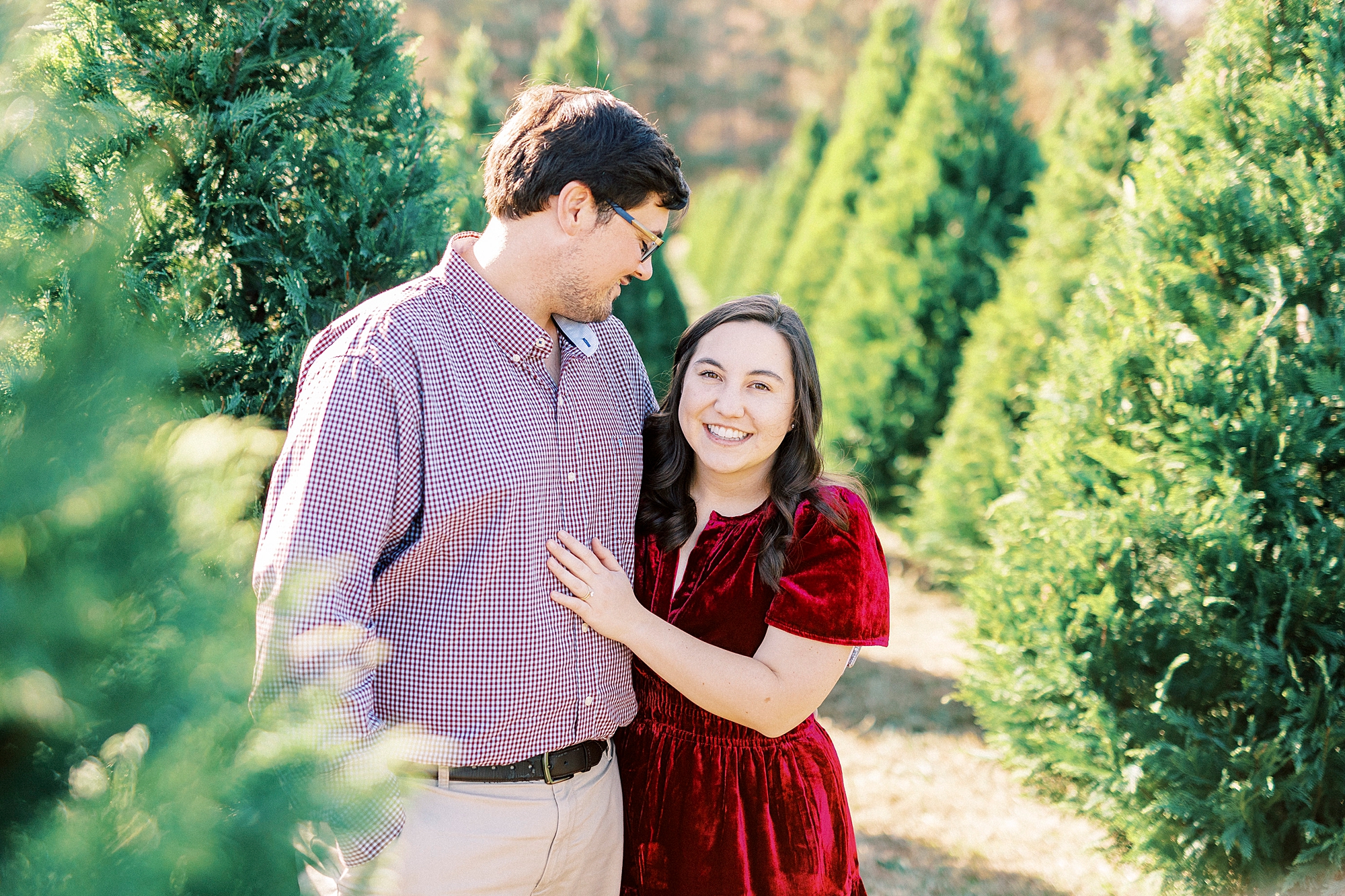 engaged couple stands between row of trees hugging during mini sessions at Penland Christmas Tree Farm