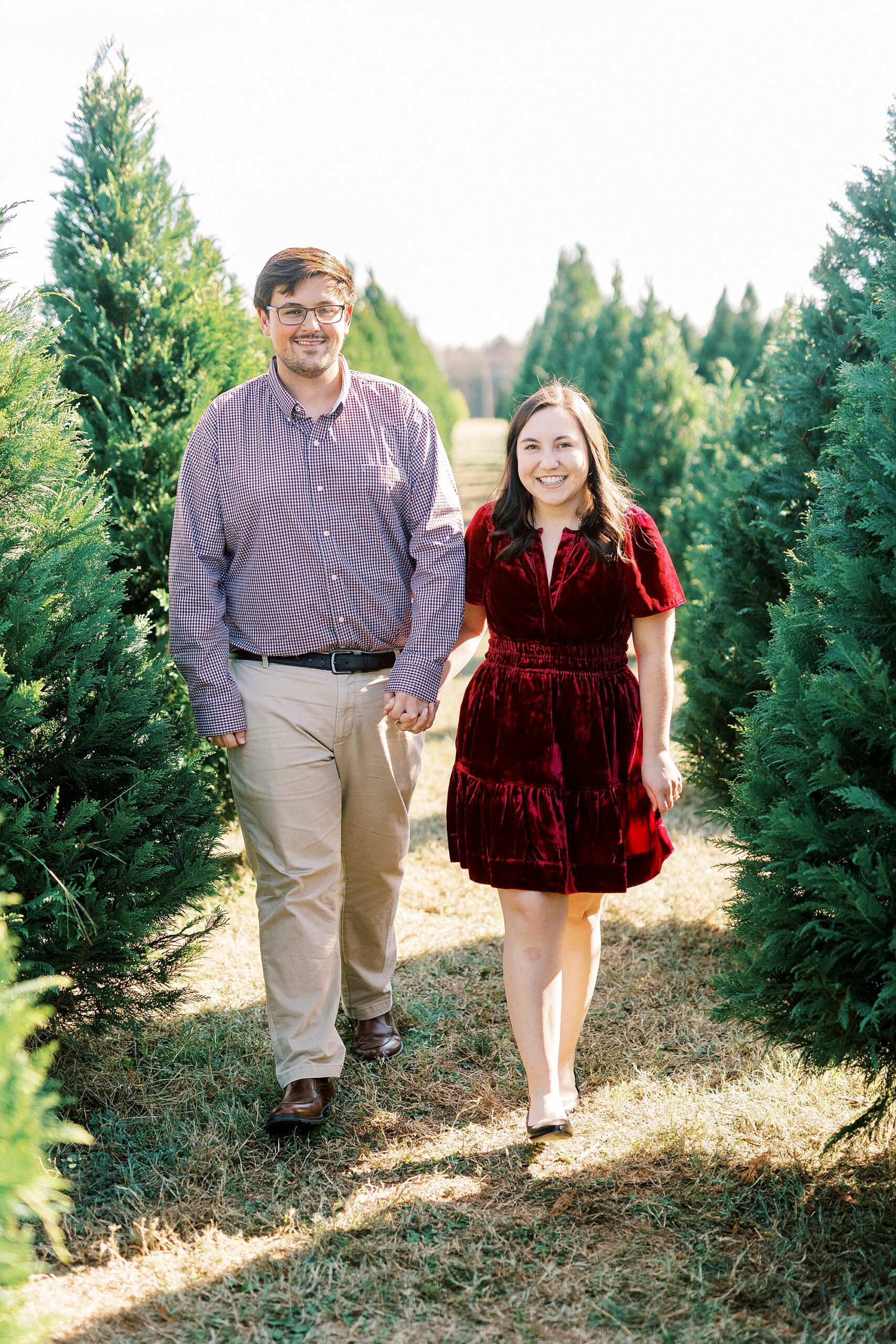 man and woman hold hands walking through row of trees during mini sessions at Penland Christmas Tree Farm