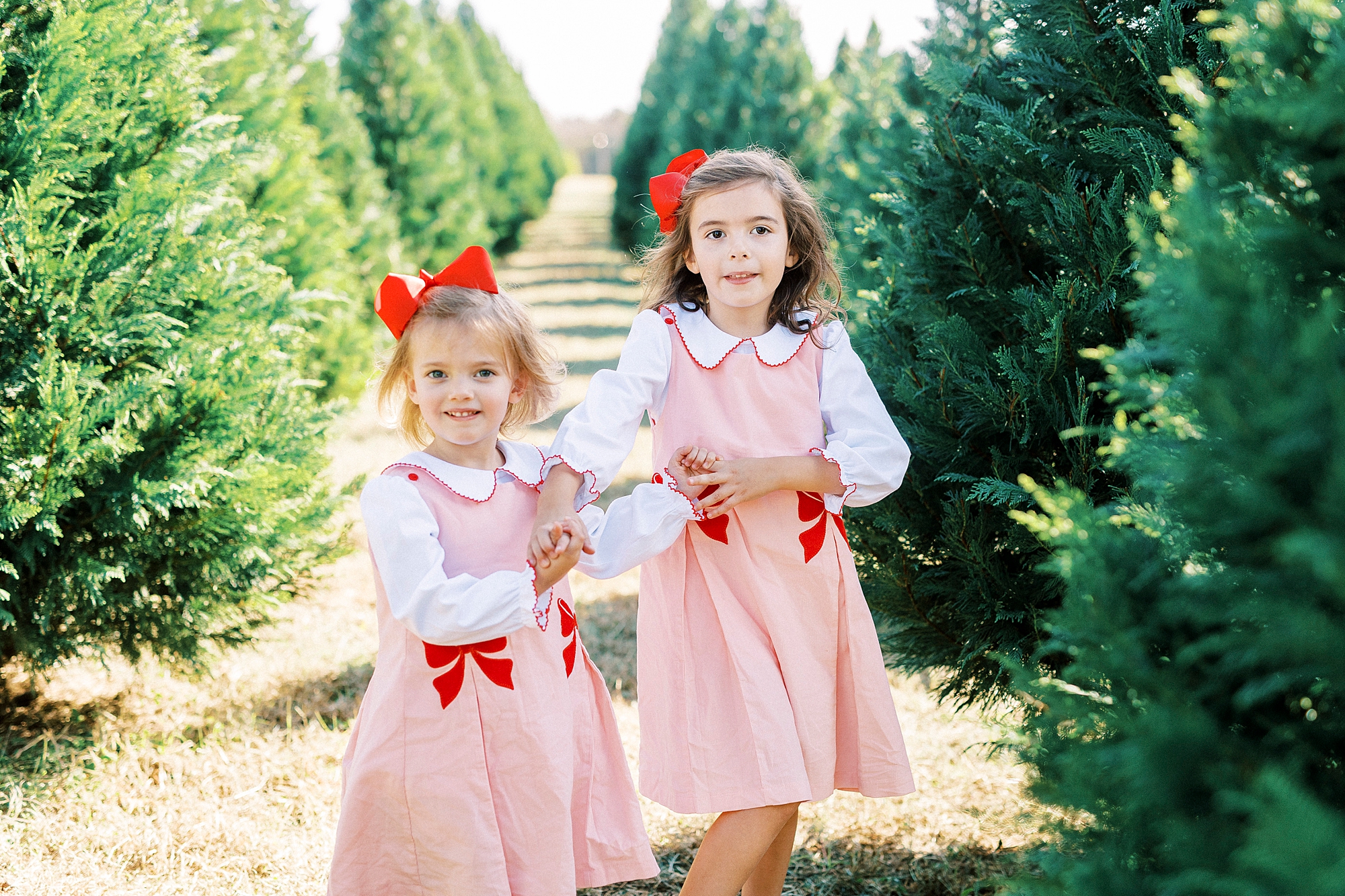 sisters in pink dresses with red bows hold hands between Christmas trees