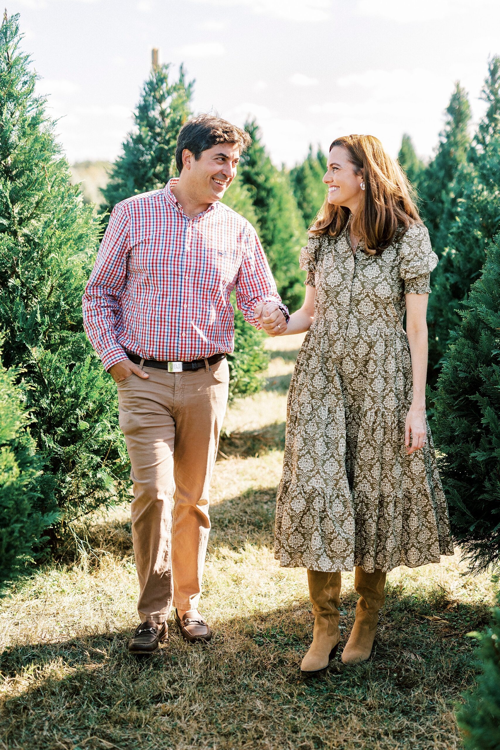 husband and wife hold hand walking between trees during mini sessions at Penland Christmas Tree Farm