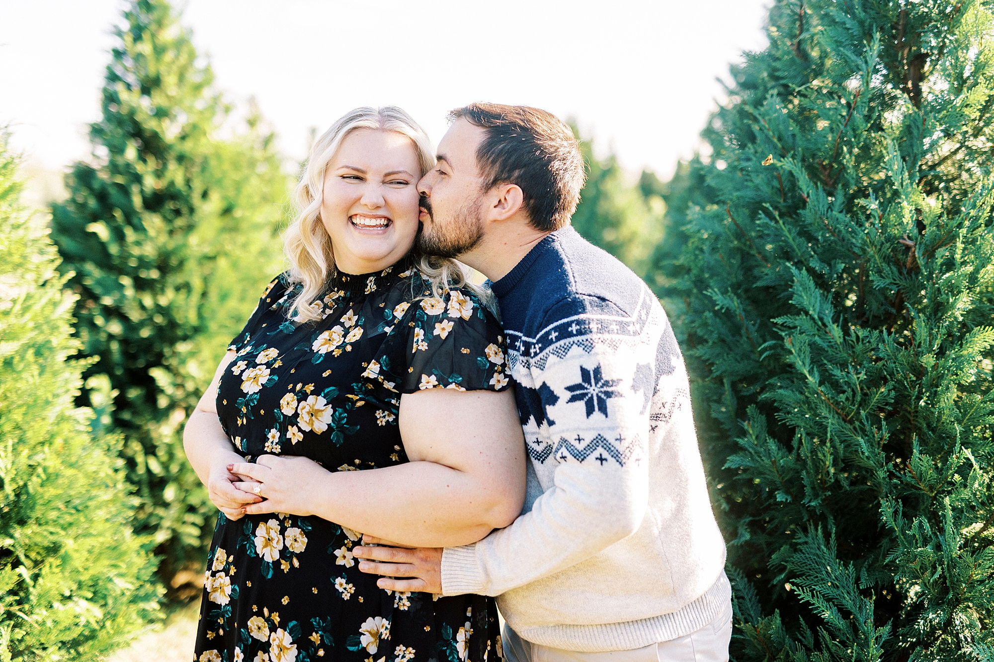 man kisses woman from behind making her smile at Christmas tree farm