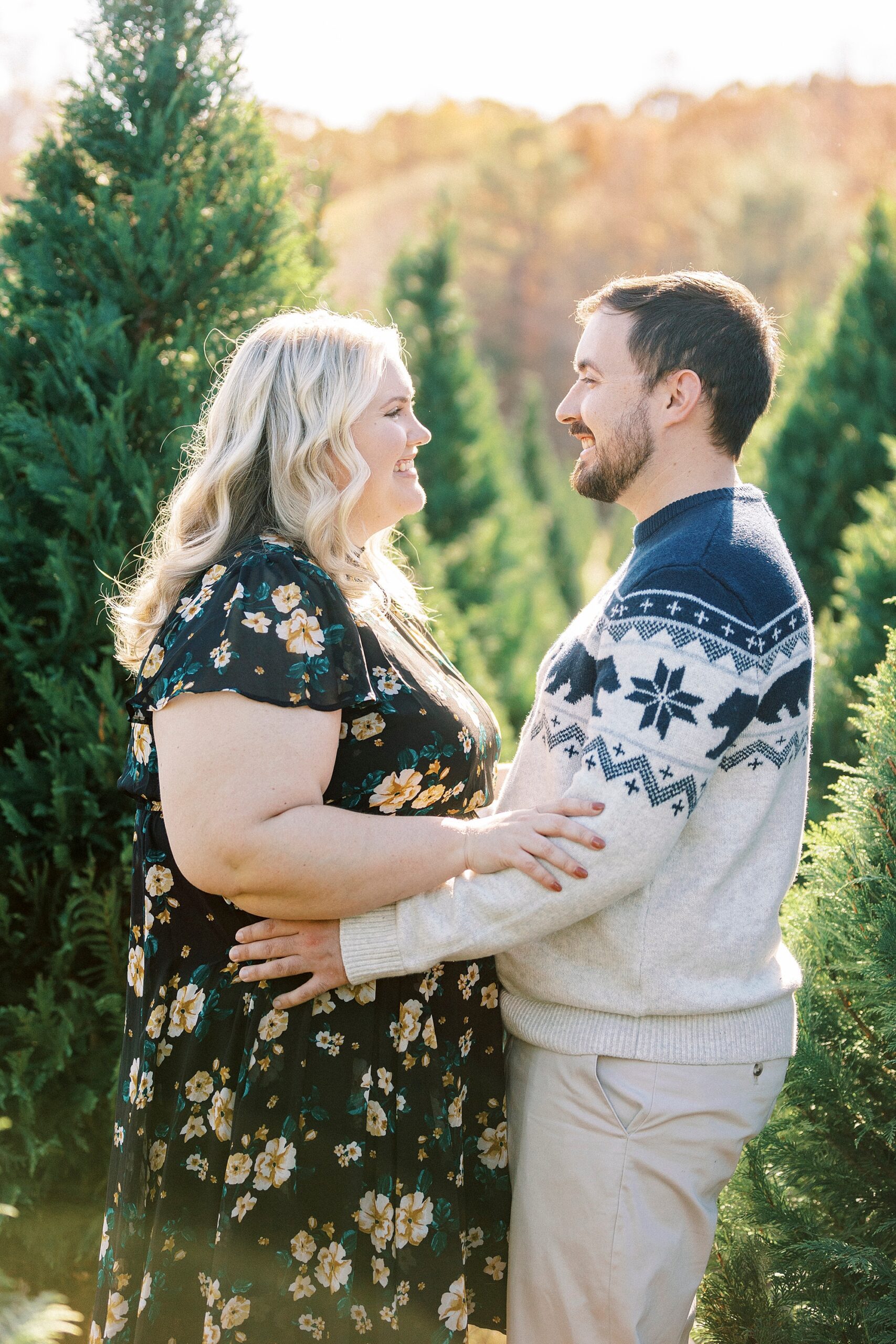 engaged couple smiles together between trees during holiday mini sessions at tree farm