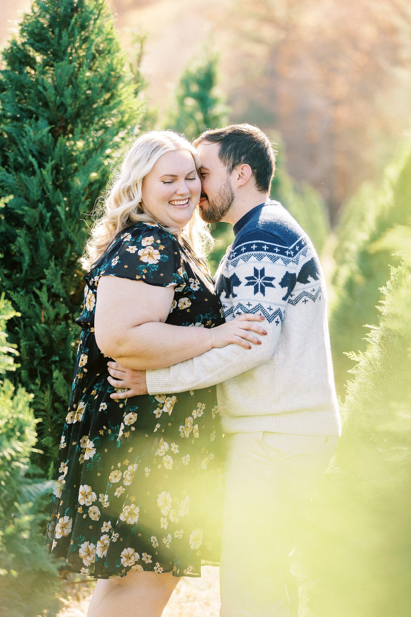 man nuzzles woman's cheek between Christmas trees during holiday mini sessions at tree farm