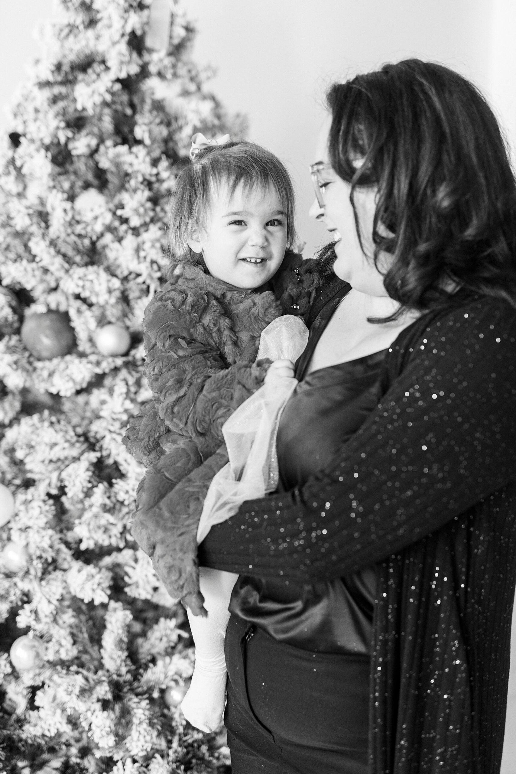 mom holds daughter on hip by tree during Christmas portraits in Downtown Concord studio