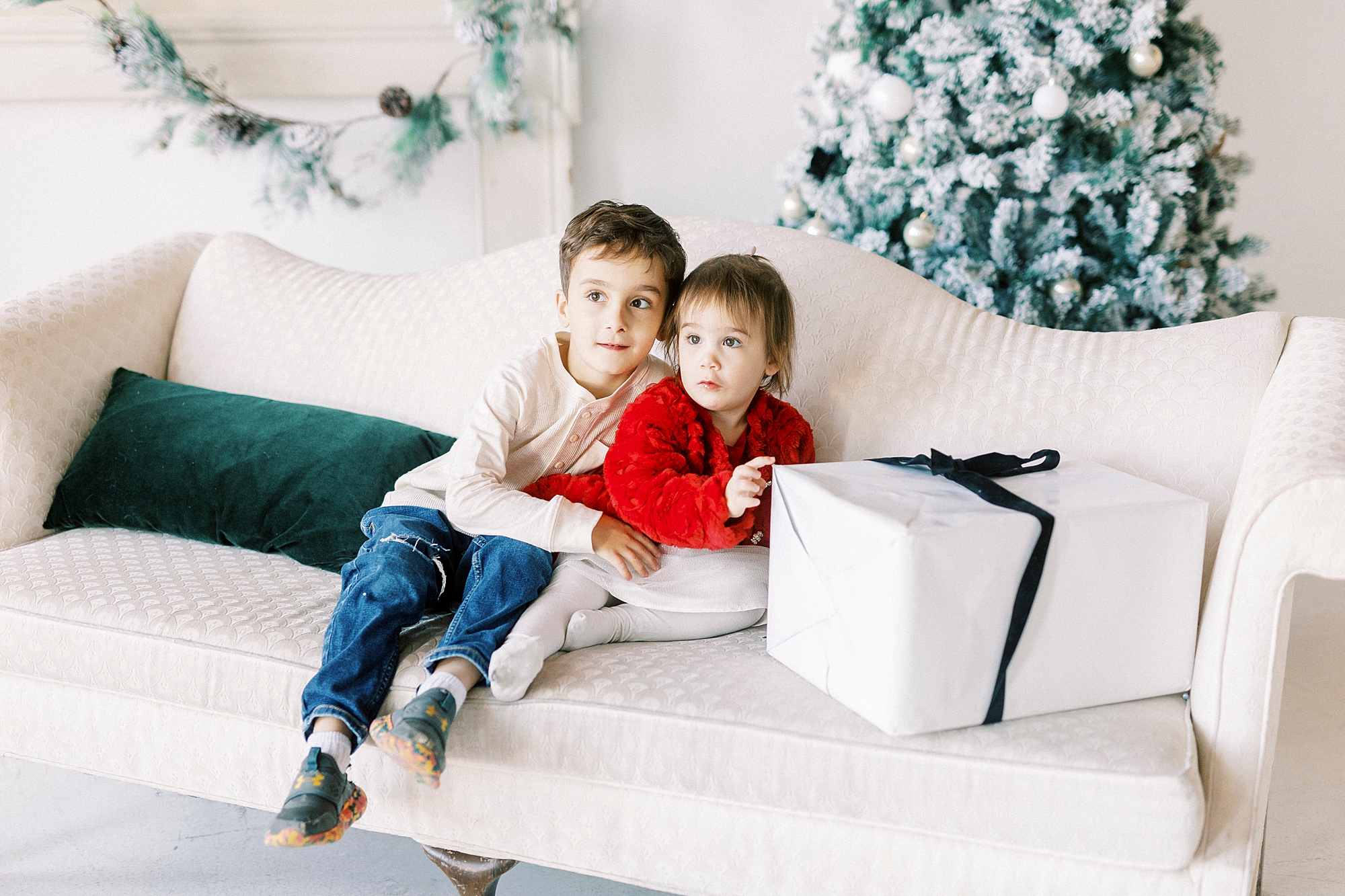 two siblings sit next to wrapped present on sofa during Christmas portraits in Downtown Concord studio