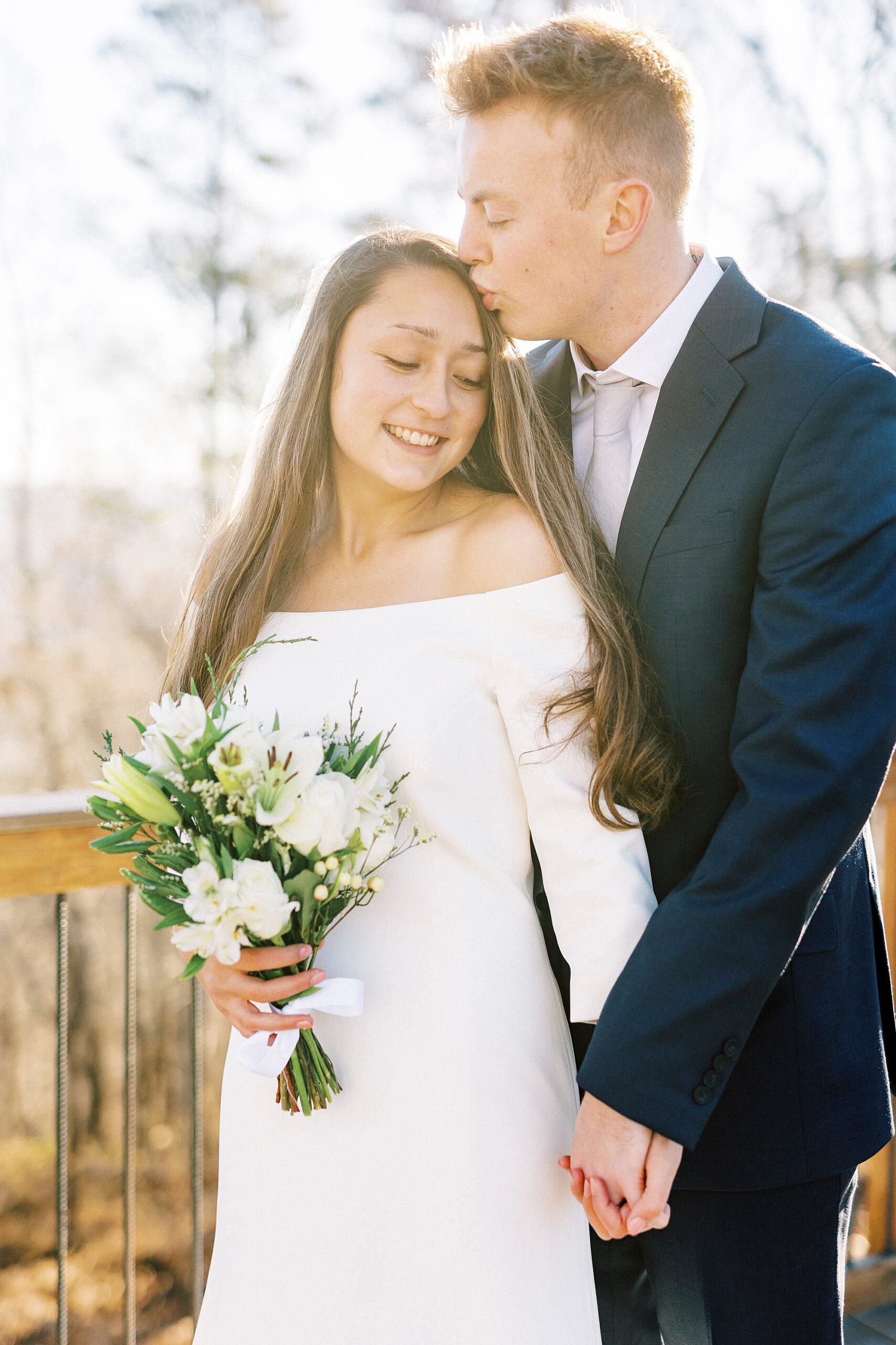 groom holds bride's hand and kisses her forehead on wooden patio at Morrow Mountain
