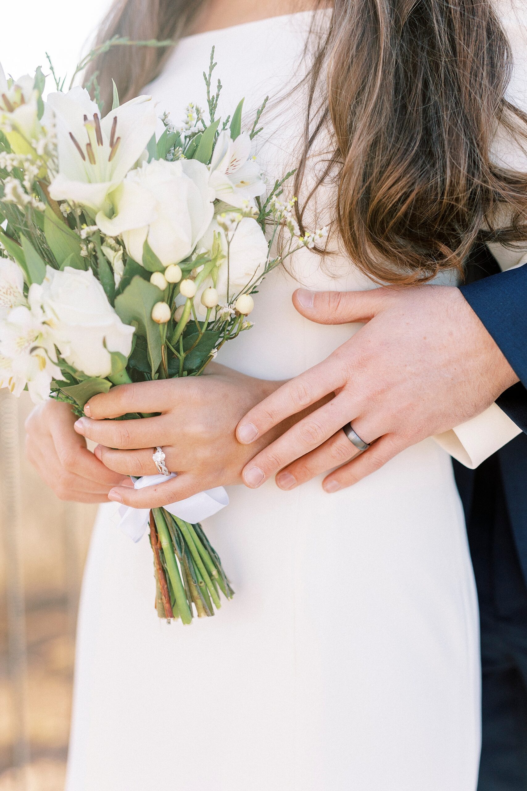 groom hugs bride from behind with bouquet of ivory flowers 