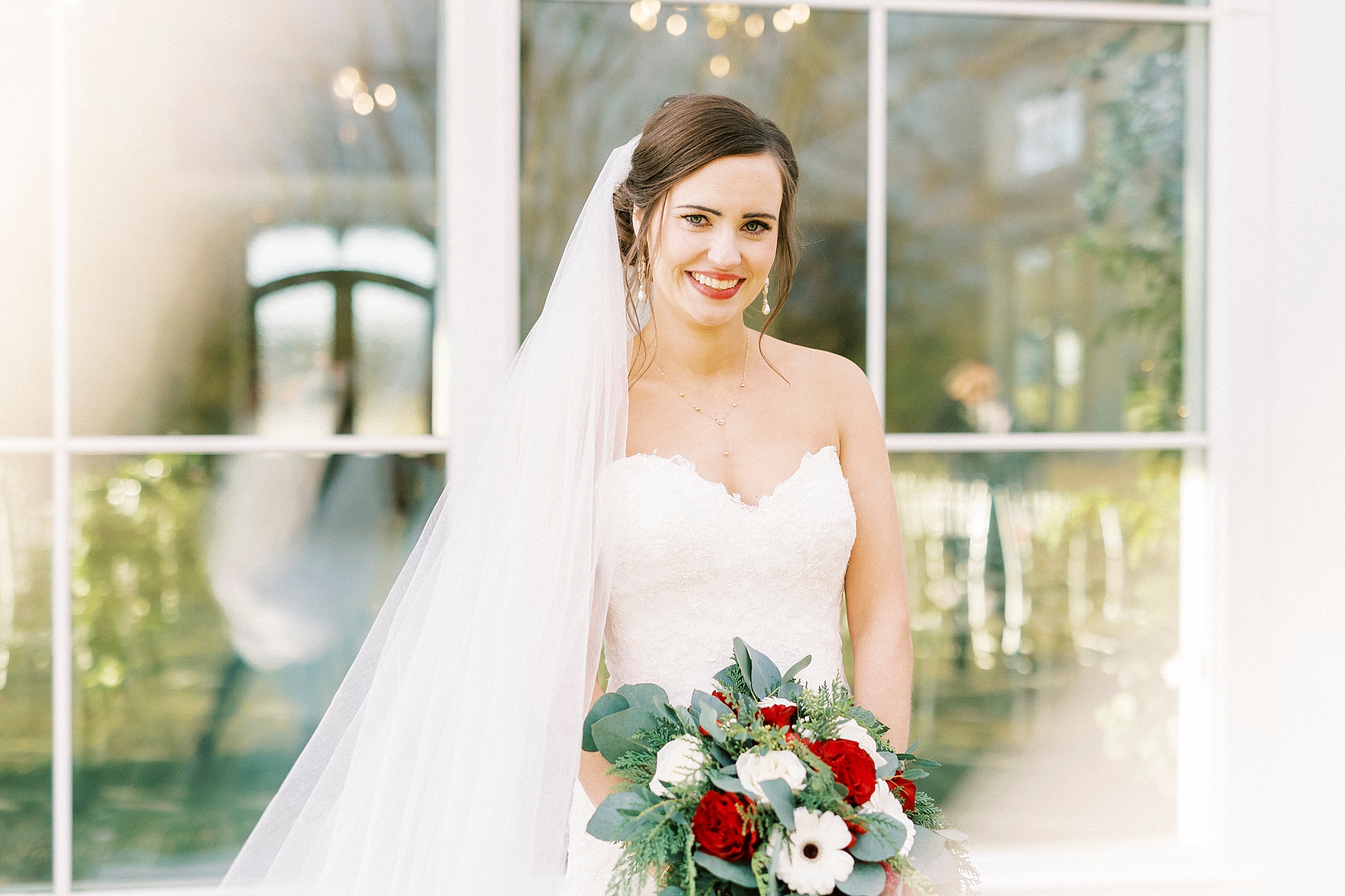 bride stands with red and white bouquet in front of chapel windows at Chickadee Hill Farms