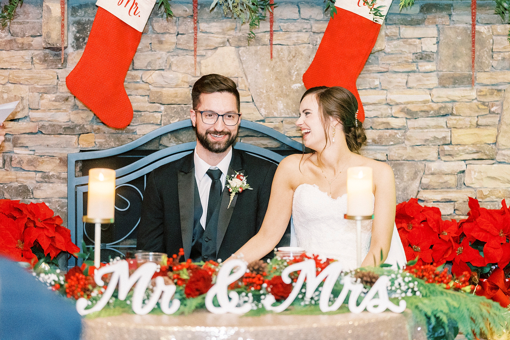 bride and groom sit at sweetheart table by stone fireplace at Chickadee Hill Farms with red stockings behind them 