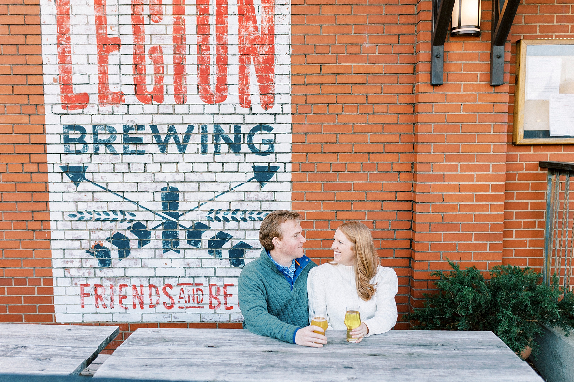 engaged couple sits at picnic table in front of brick wall at Legion Brewing in Charlotte NC