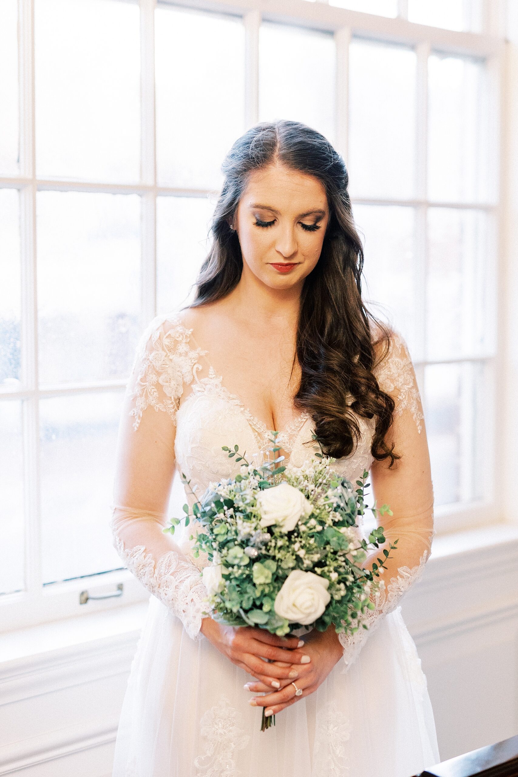 bride smiles down at bouquet of white roses and greenery in front of church window
