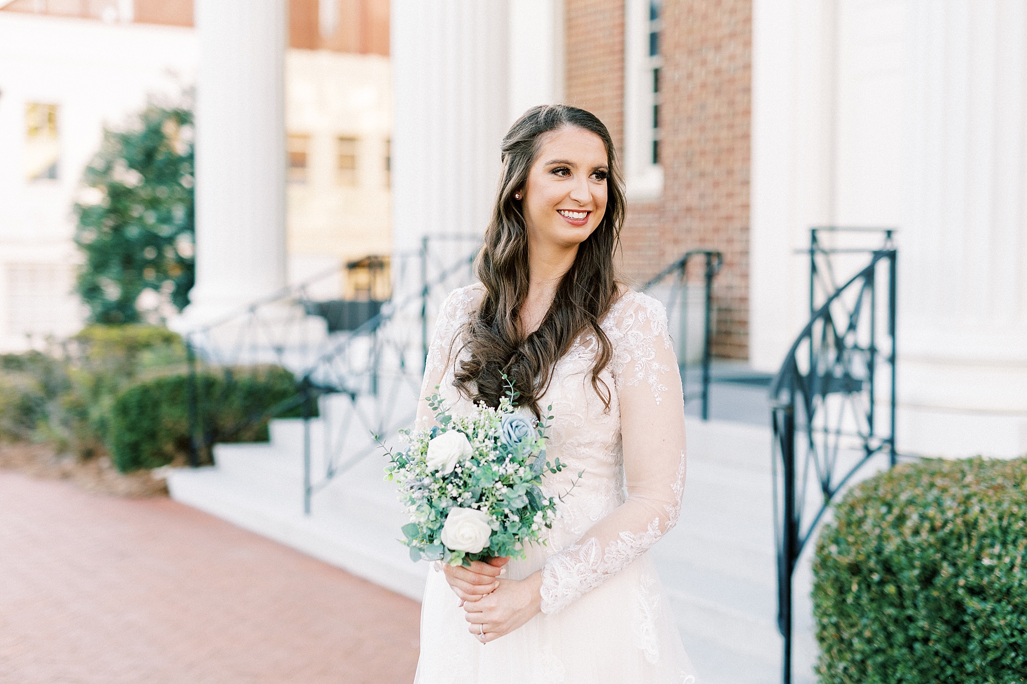 bride smiles holding bouquet of white flowers outside Downtown Kannapolis church
