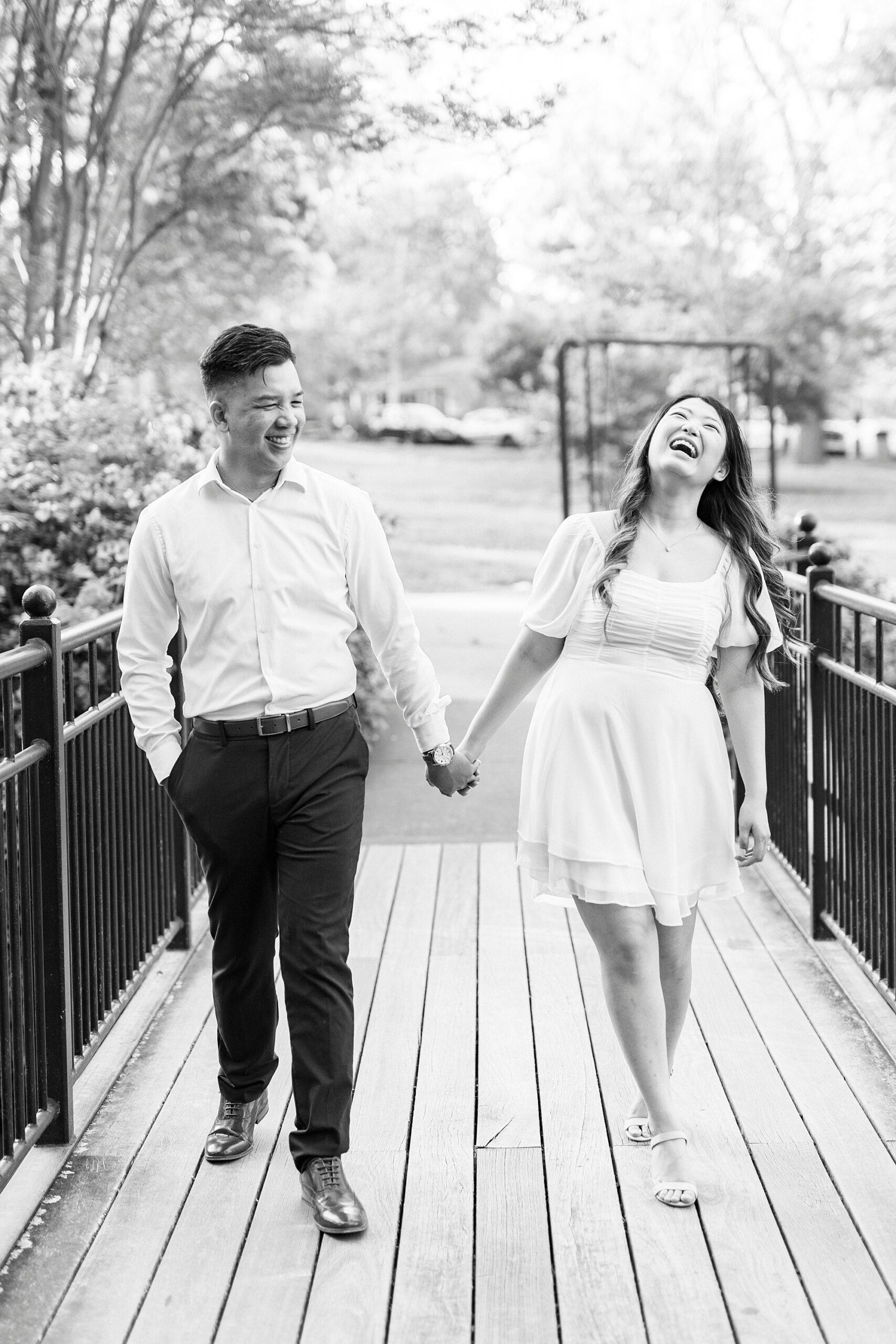 engaged couple holds hands while woman laughs walking on wooden bridge 