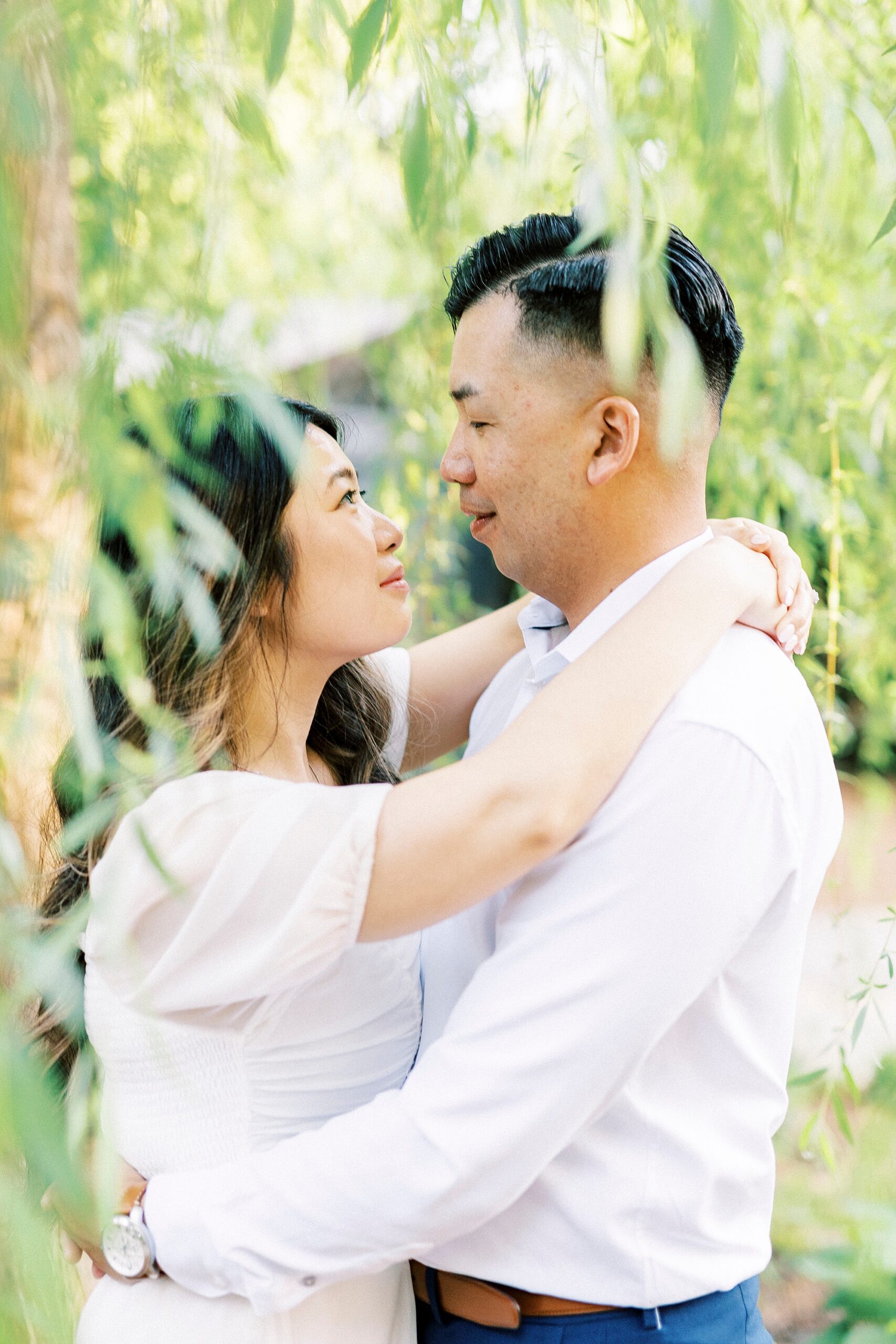 couple hugs among leaves of weeping willow smiling at each other