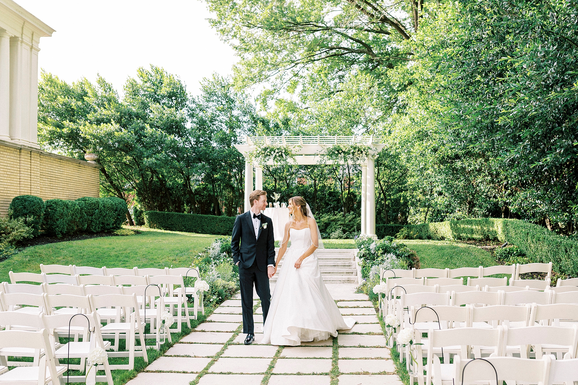 bride and groom hold hands walking between white chairs on lawn