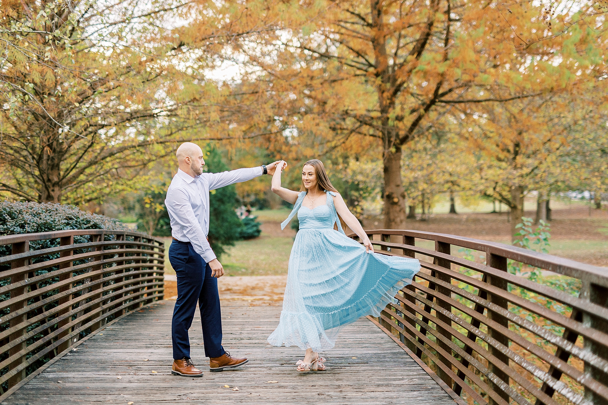 man twirls woman in light blue dress on wooden bridge inside Glencairn Garden