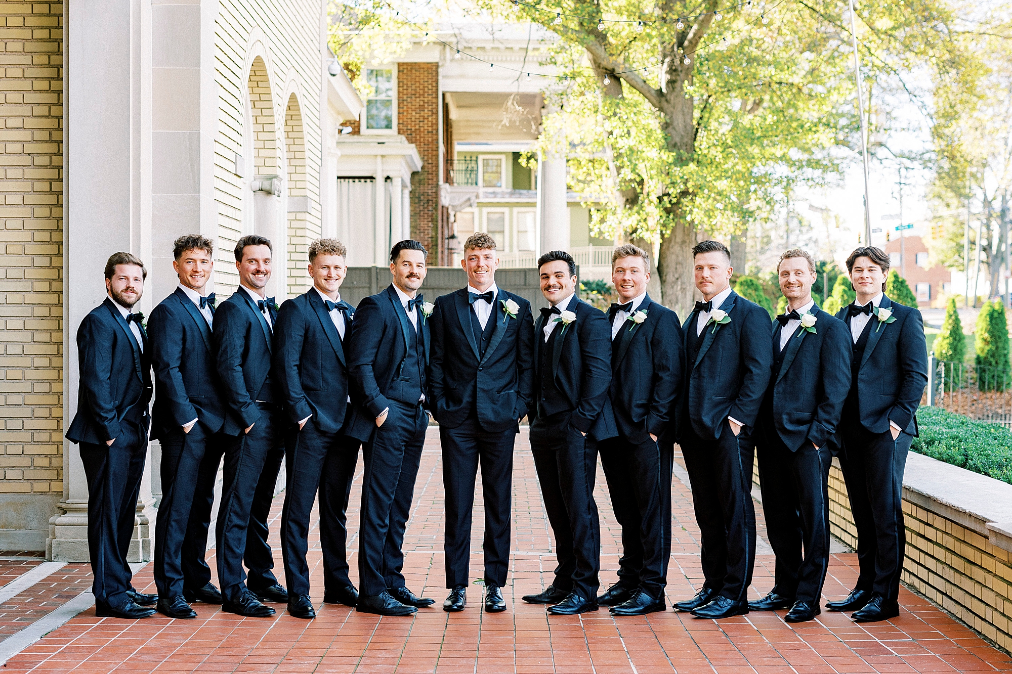 groom stands with groomsmen in a line on brick patio at v