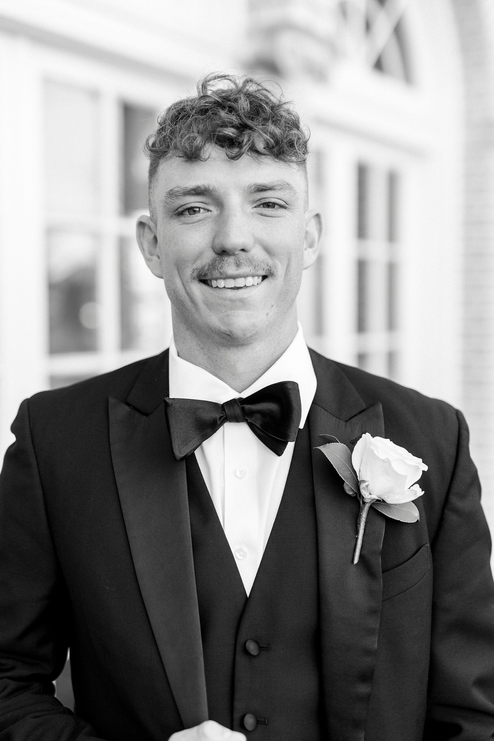 groom in suit stands smiling with white rose boutineere