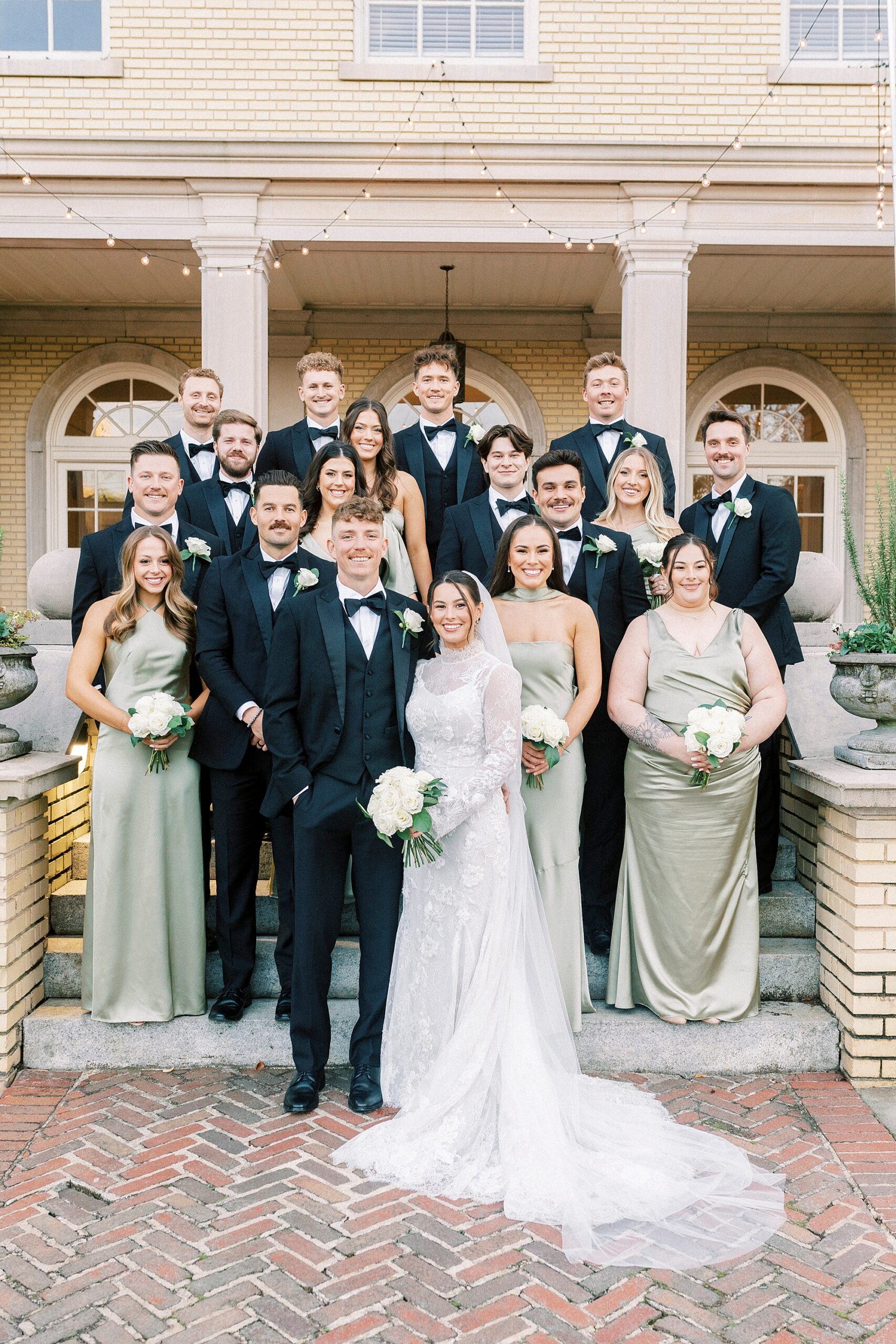 nelwwyds stand on steps of Separk Mansion with wedding party in sage green and black attire