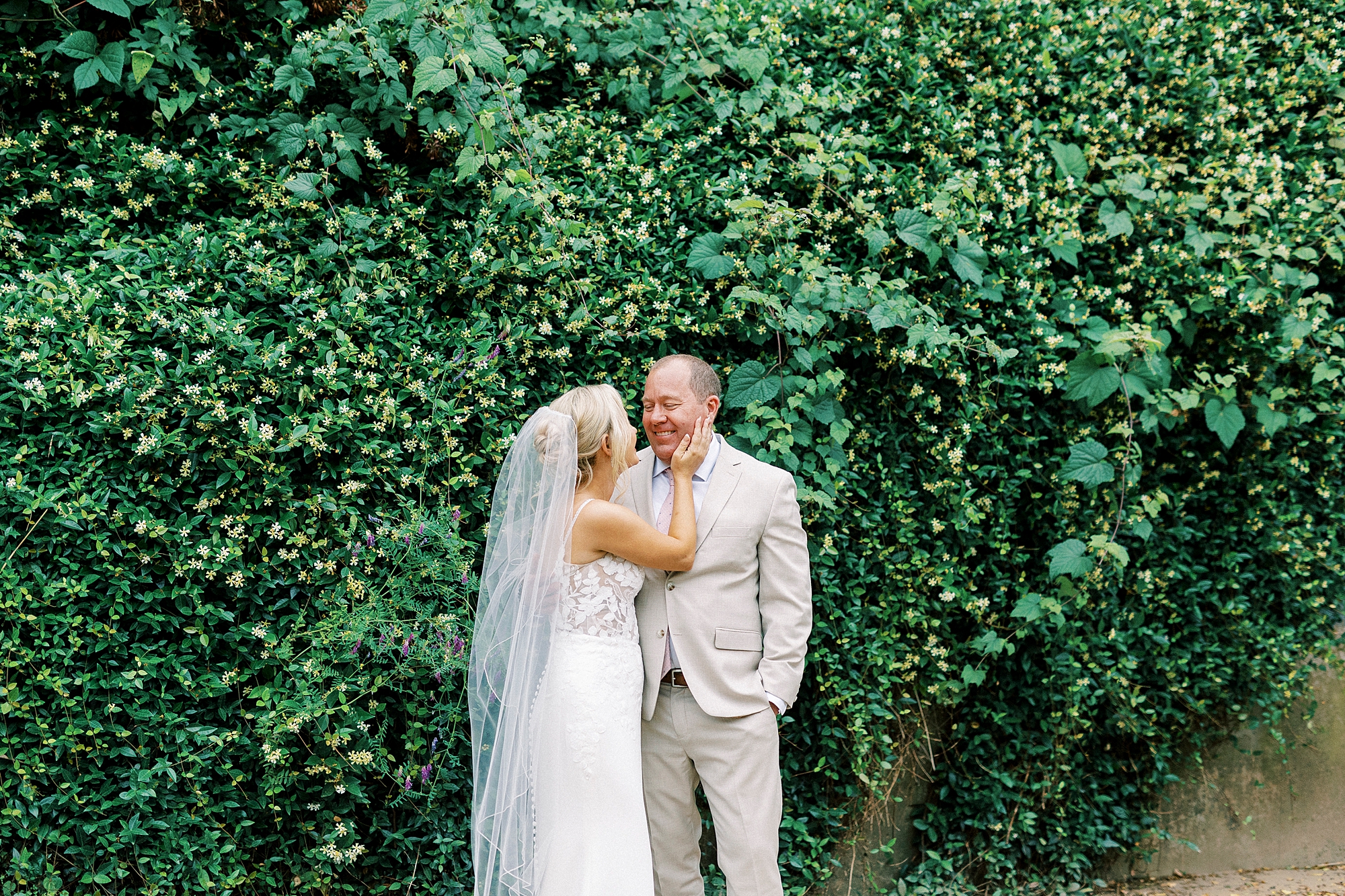 bride smiles up at groom in front of green bushes