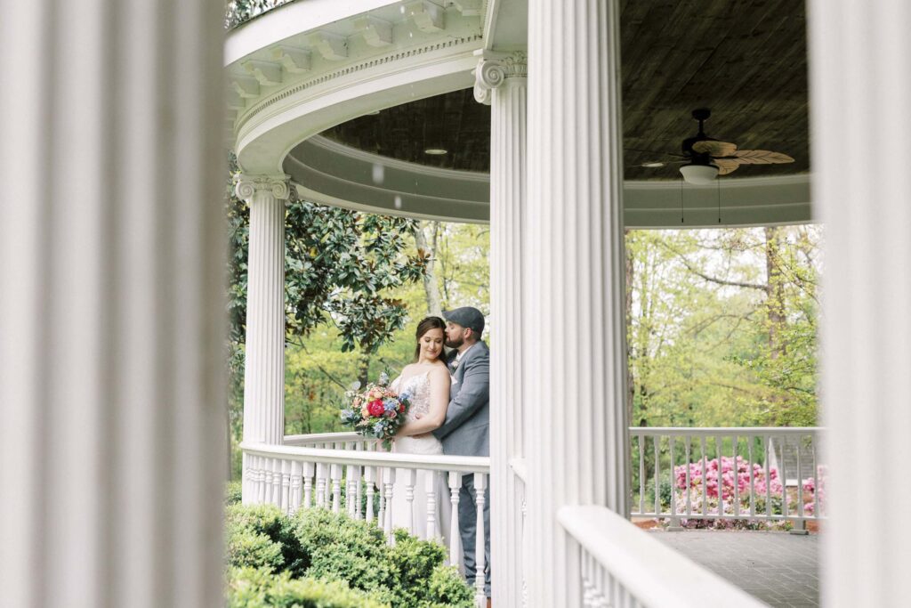 groom kisses bride on forehead on porch of historic home at Saratoga springs wedding venue in mount pleasant North Carolina